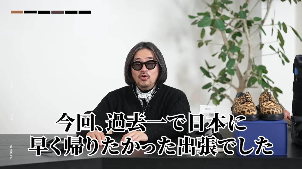 机の前で身振りを交えながら話すサングラス姿の男性