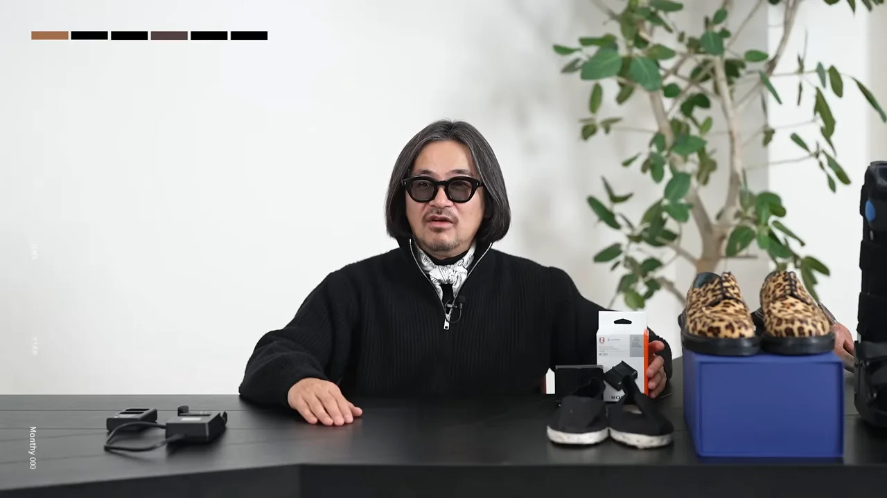 机の上に靴や小物を並べて正面を向いて話す男性