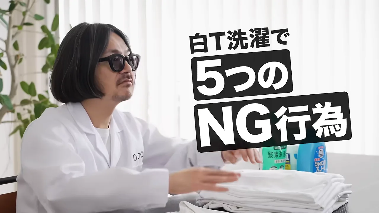 白衣とサングラスを身につけた男性が、机の上に積まれた白い衣類や洗剤の前に座って話している様子。