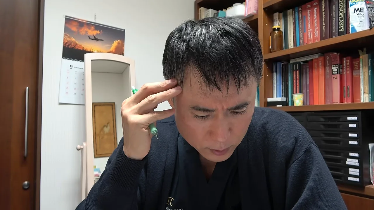 本棚を背景に座る男性がペンを持ちながらこめかみに手を当てている様子
