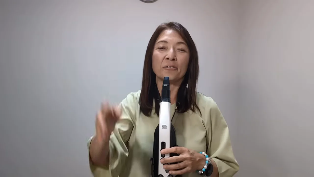 電子管楽器を手に持ち、手振りをつけて話す女性の様子。