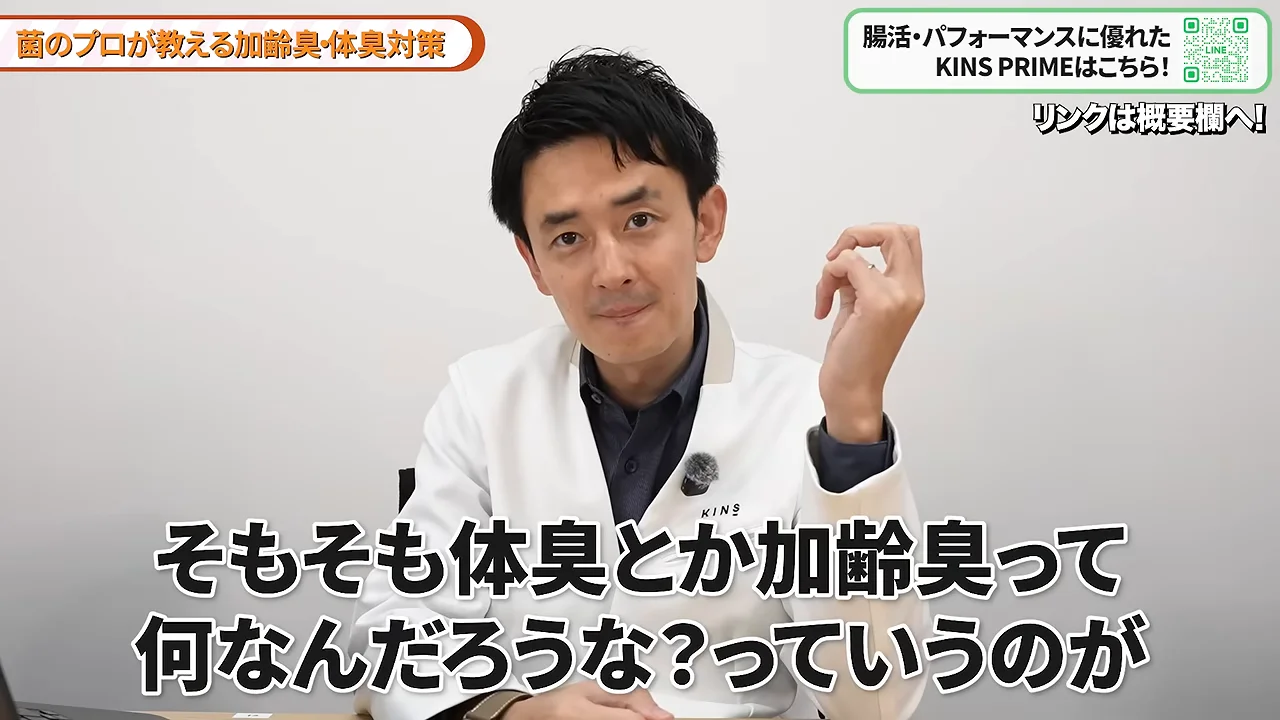 白衣を着た男性が左手を上げてカメラに向かって話している様子。