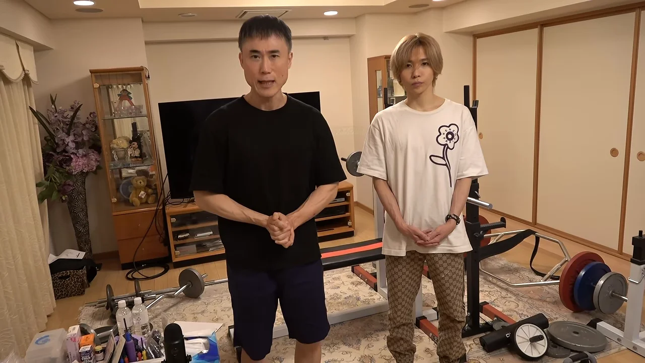 トレーニング器具が置かれた部屋で、黒いTシャツの男性と白いTシャツの男性が並んで立っています。