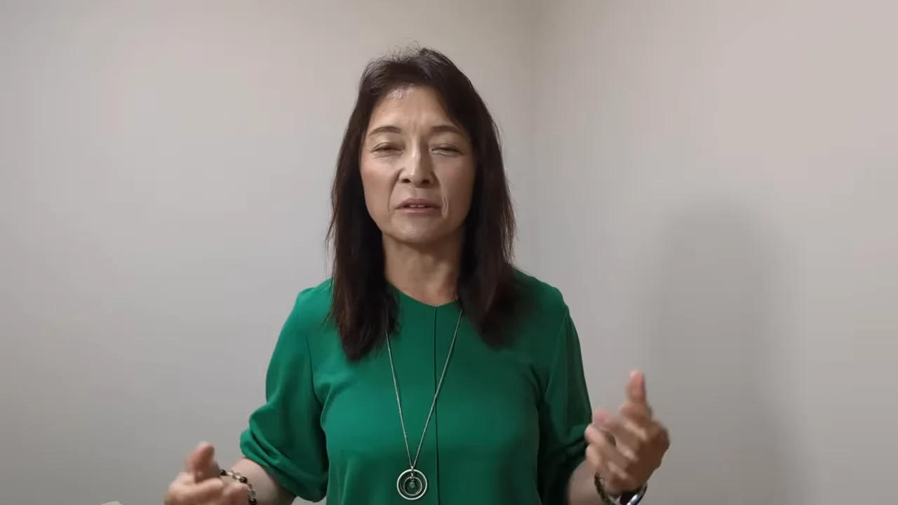 白い壁を背景に、緑色の服を着た女性が手ぶりを交えながら話しています。