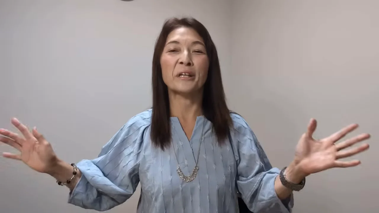 水色の服を着た女性が両手を広げてカメラに向かって話している様子。