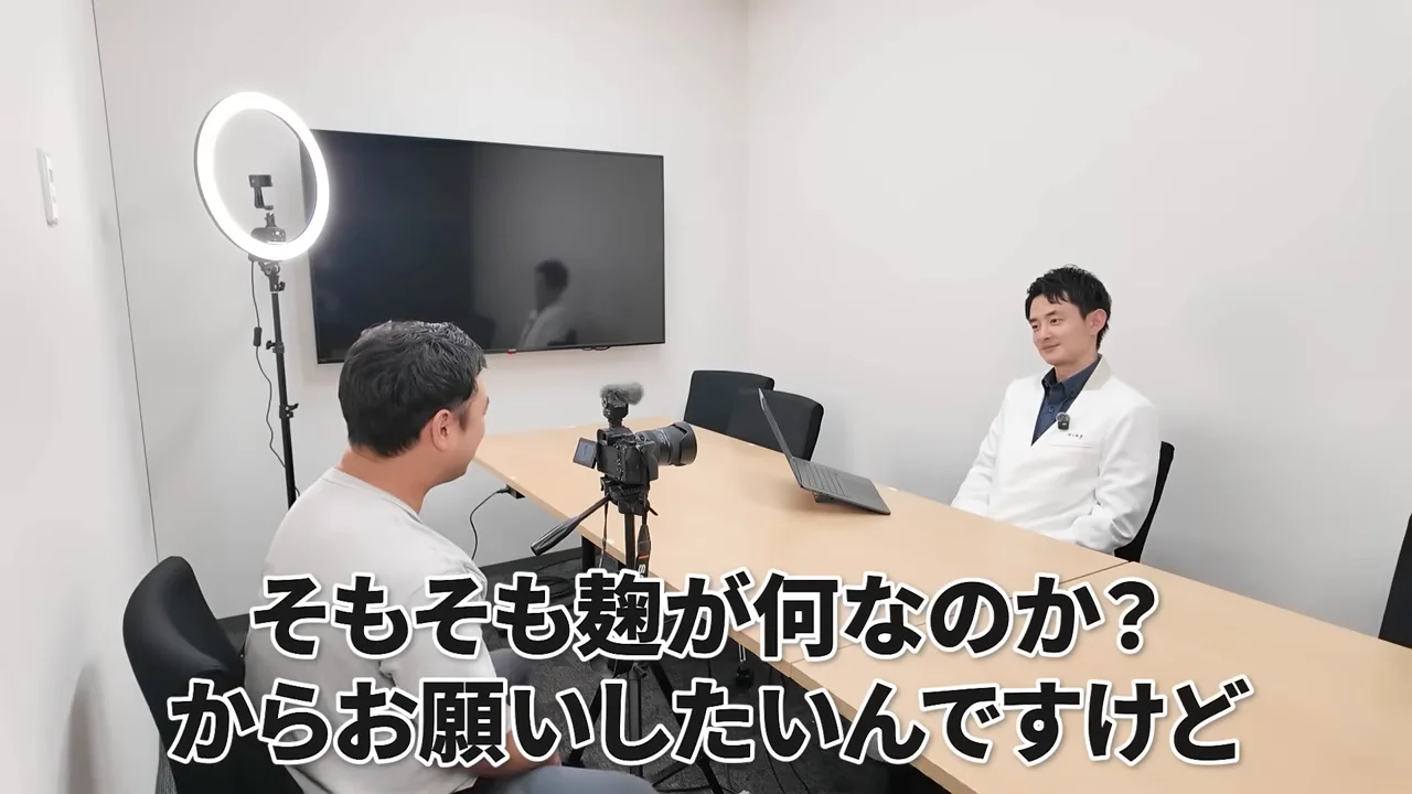 会議室でテーブルを挟んで男性2人が座り、カメラを向けて対談している様子。