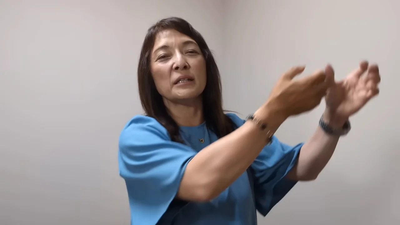 青い服を着た女性が両手を前方に掲げながら話している様子。
