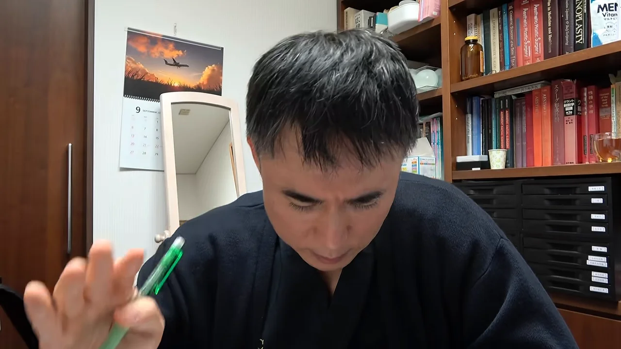 本棚を背にして座った男性が、緑色のペンを手に持ち下を向いている様子。