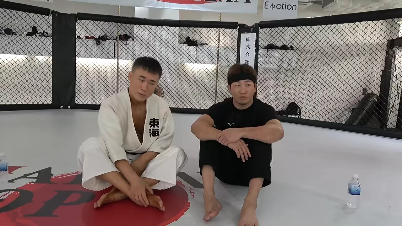 柔道着を着た男性と黒い服の男性が、リラックスした様子でマットの上に座っています。