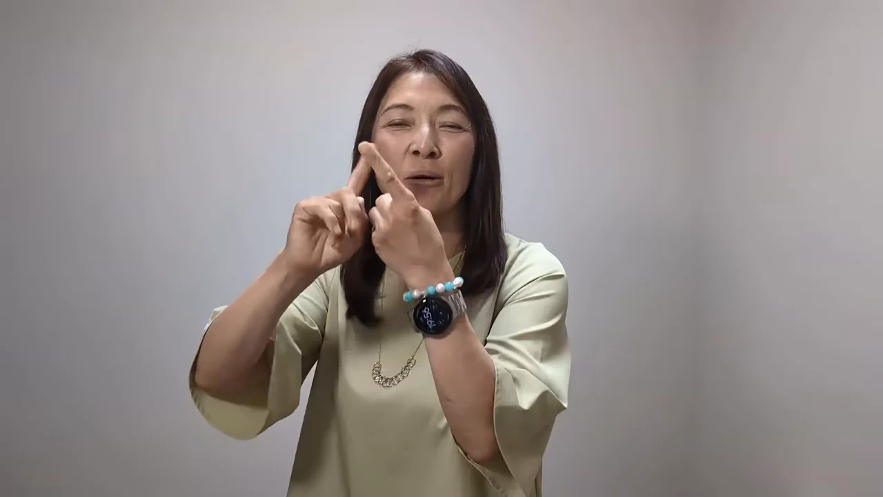 薄緑色の服を着た女性が、両手の人差し指を立てて合わせる仕草をしながら話しています。
