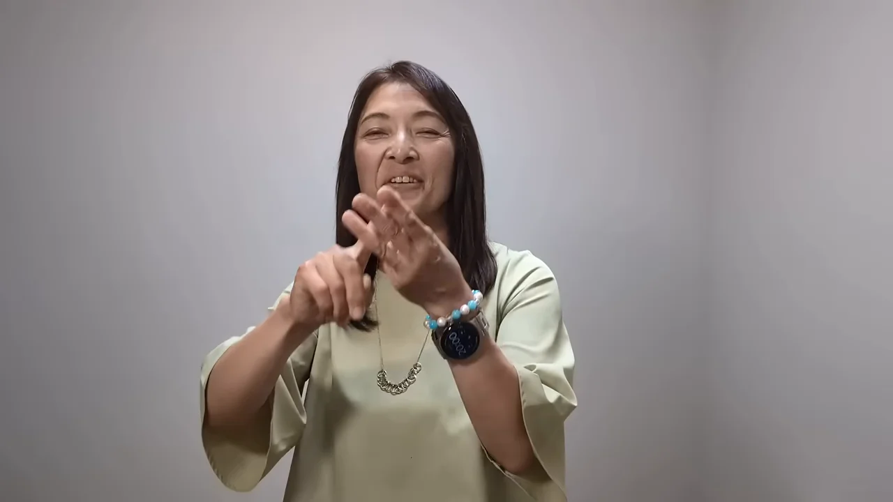 薄緑色の服を着た女性が、手元で指を重ねるジェスチャーをしながら笑顔で話しています。
