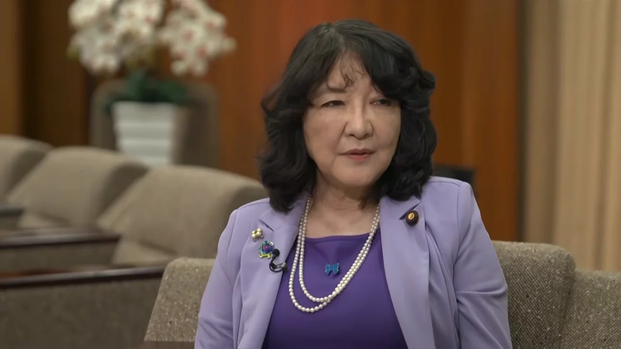 薄紫色のジャケットを着用した女性が、椅子に座って落ち着いた様子で話しています。