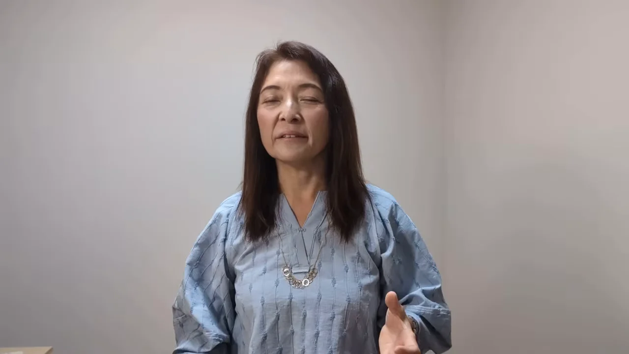 白い壁の前で青い服を着た女性がカメラに向かって話しています。