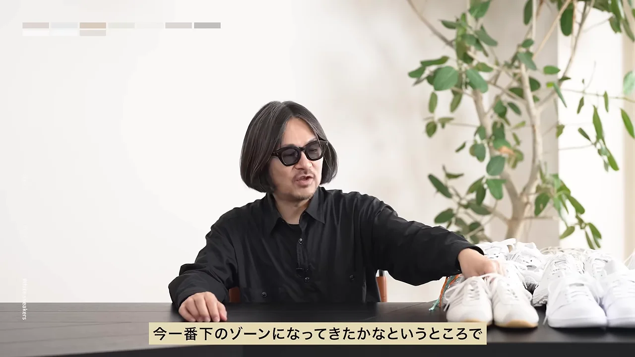 黒いシャツを着てサングラスをかけた人物が、テーブルの上に並べられた白いスニーカーに手を伸ばして話しています。