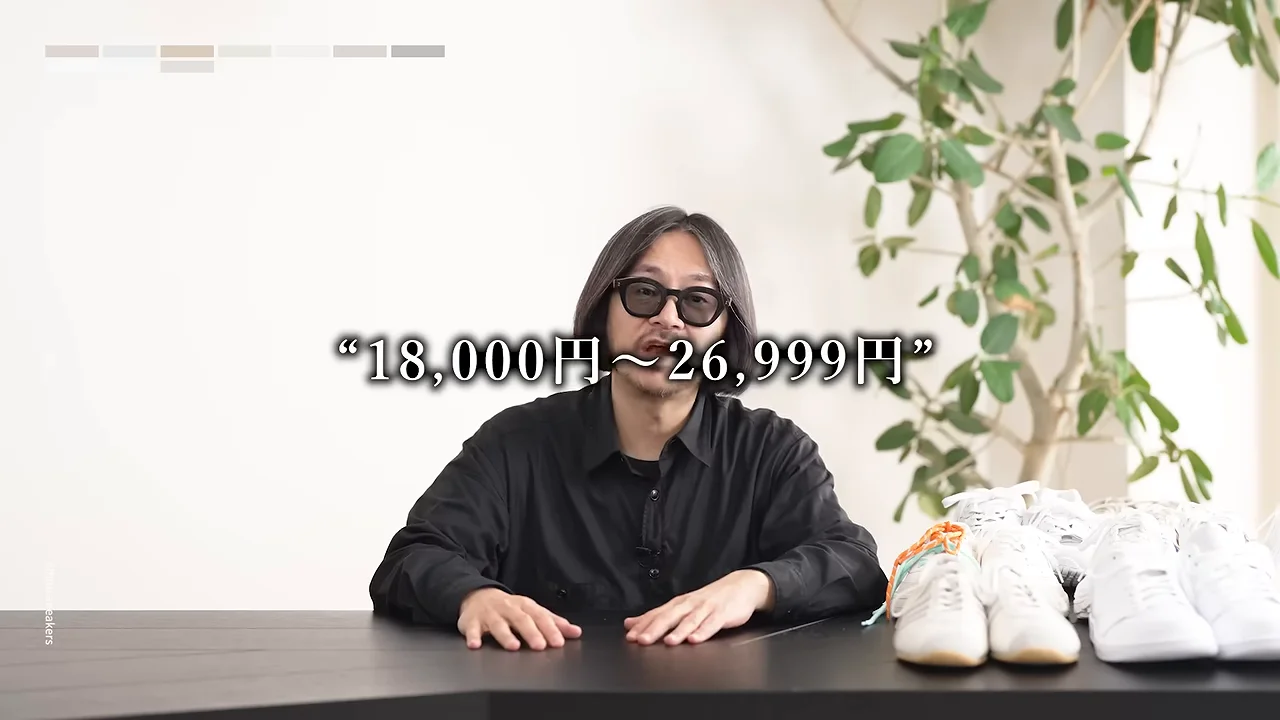 テーブルの上に複数の白いスニーカーが並べられており、その前に座る人物の前に金額のテキストが表示されています。