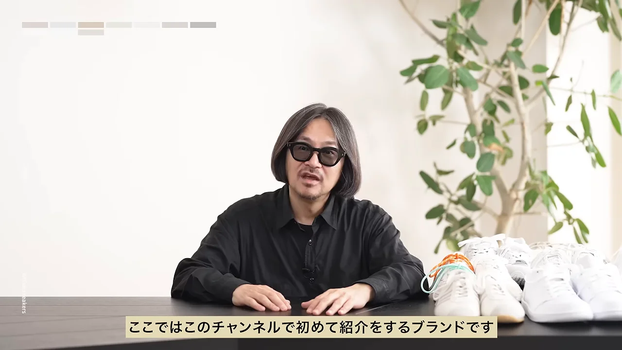 サングラスをかけた人物が、白いスニーカーが並ぶテーブルの前に座り正面を向いて話しています。