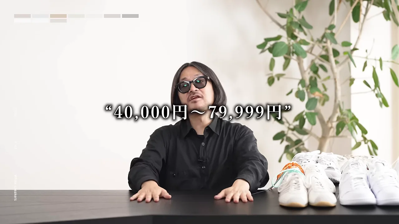 黒いシャツを着た人物が、白いスニーカーが置かれたテーブルに両手を広げて座り、画面中央にテキストが表示されています。