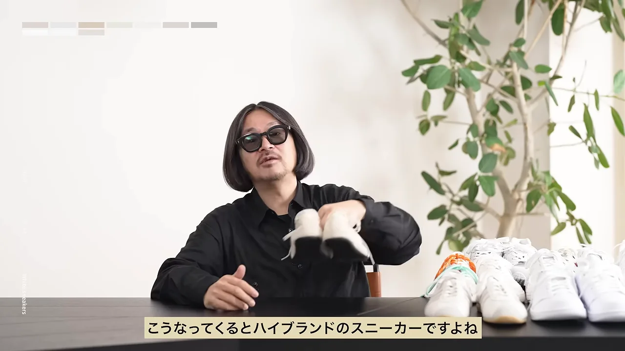 サングラスをかけた人物が、テーブルの前で白い靴を一足両手で持ち上げています。