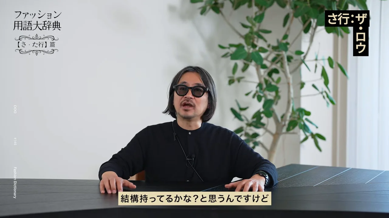黒い服とサングラスを身につけた男性がテーブルの前に座って話している様子。
