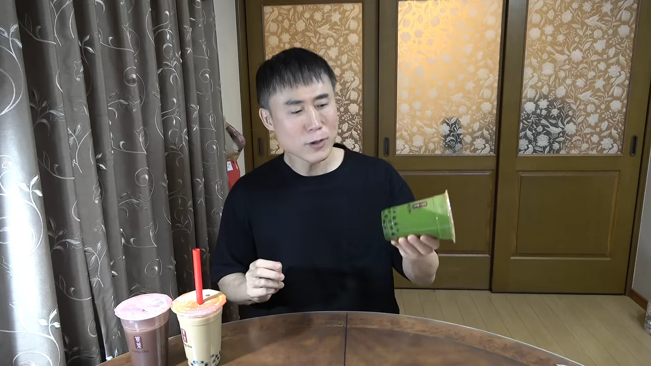 男性が抹茶ミルクティーのカップを持ち上げてカメラに見せている場面。