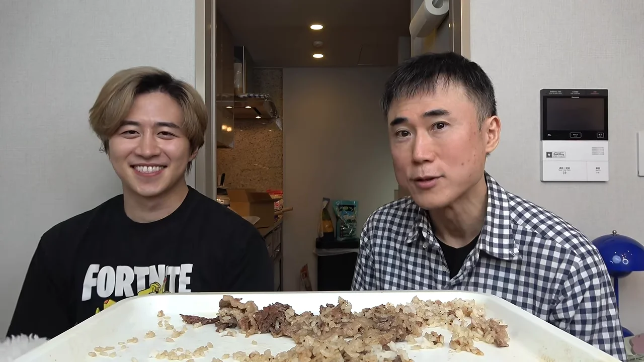 料理を前にしたトレイを挟んで並び、カメラに向かって話したり笑顔を見せたりする2人の男性。