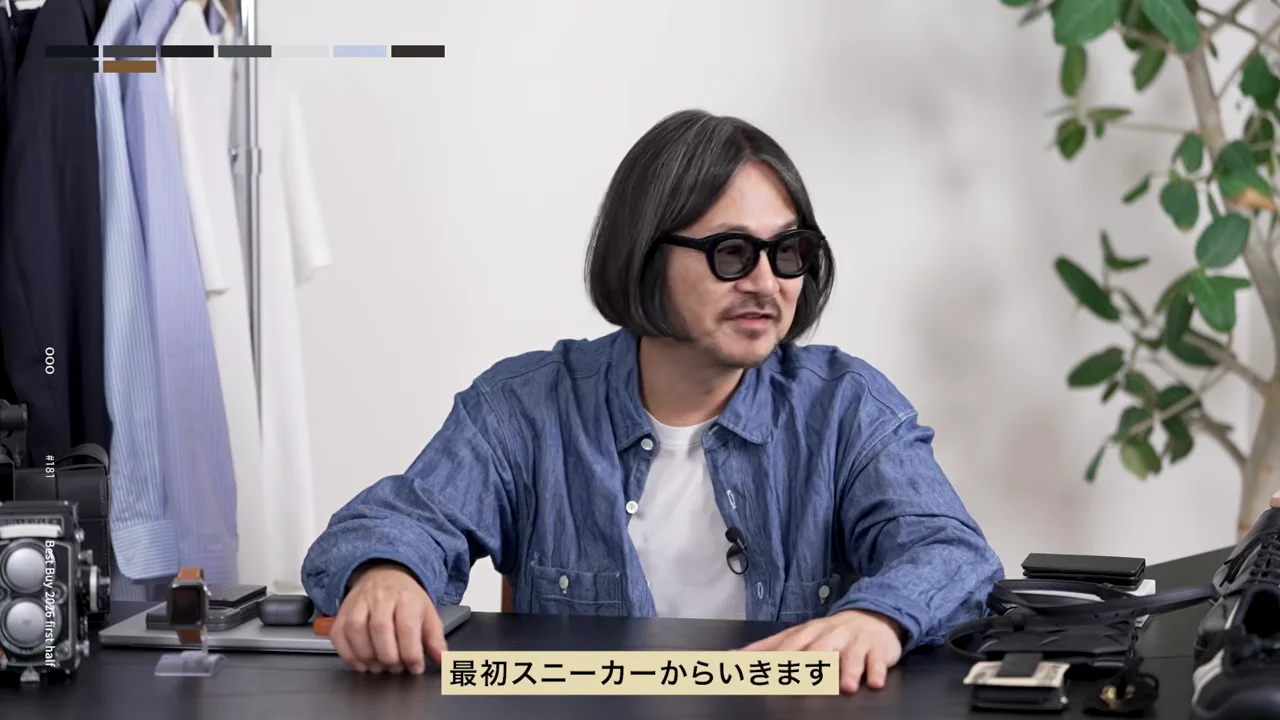眼鏡をかけた男性が、衣類や靴の並ぶテーブルに両手を置いて座っている。