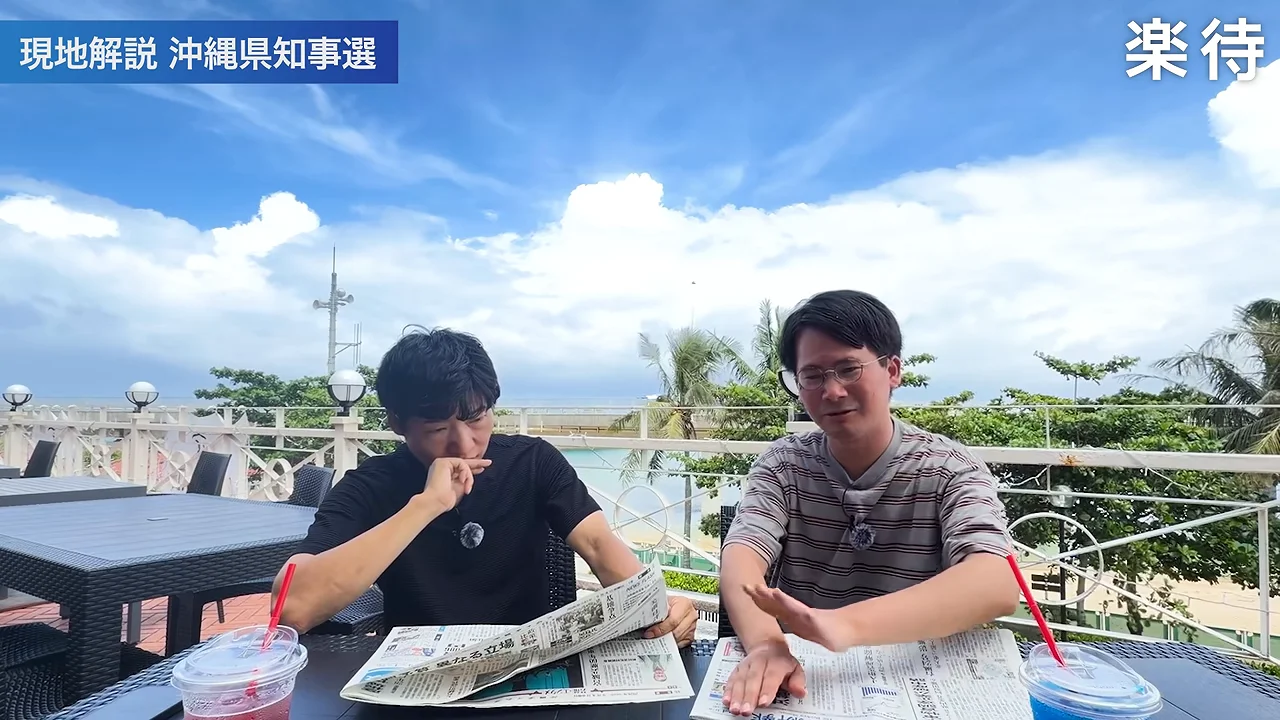 テーブルの上に新聞を広げて話す2人の男性。