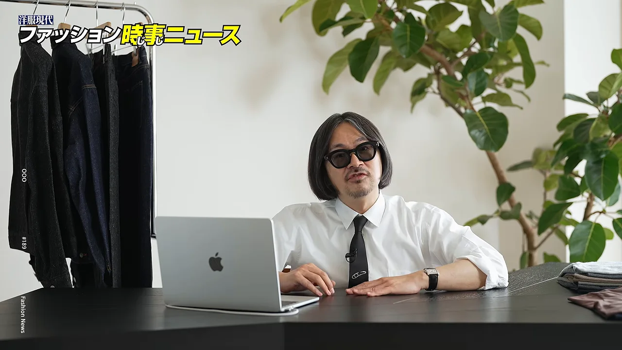 服が掛かったラックを背に、机にノートパソコンを置いた男性が正面を向いています。