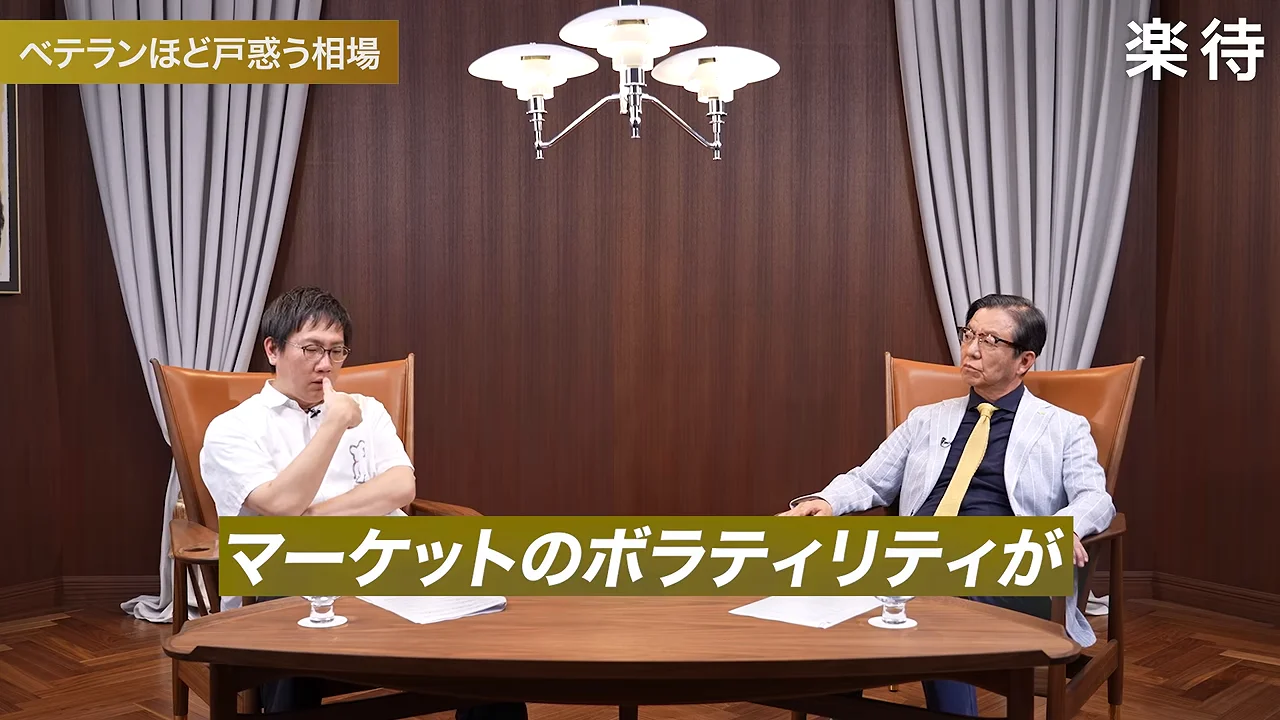 テーブルを挟んで座る2人の男性のうち、左側の男性が口元に指を当てている様子。