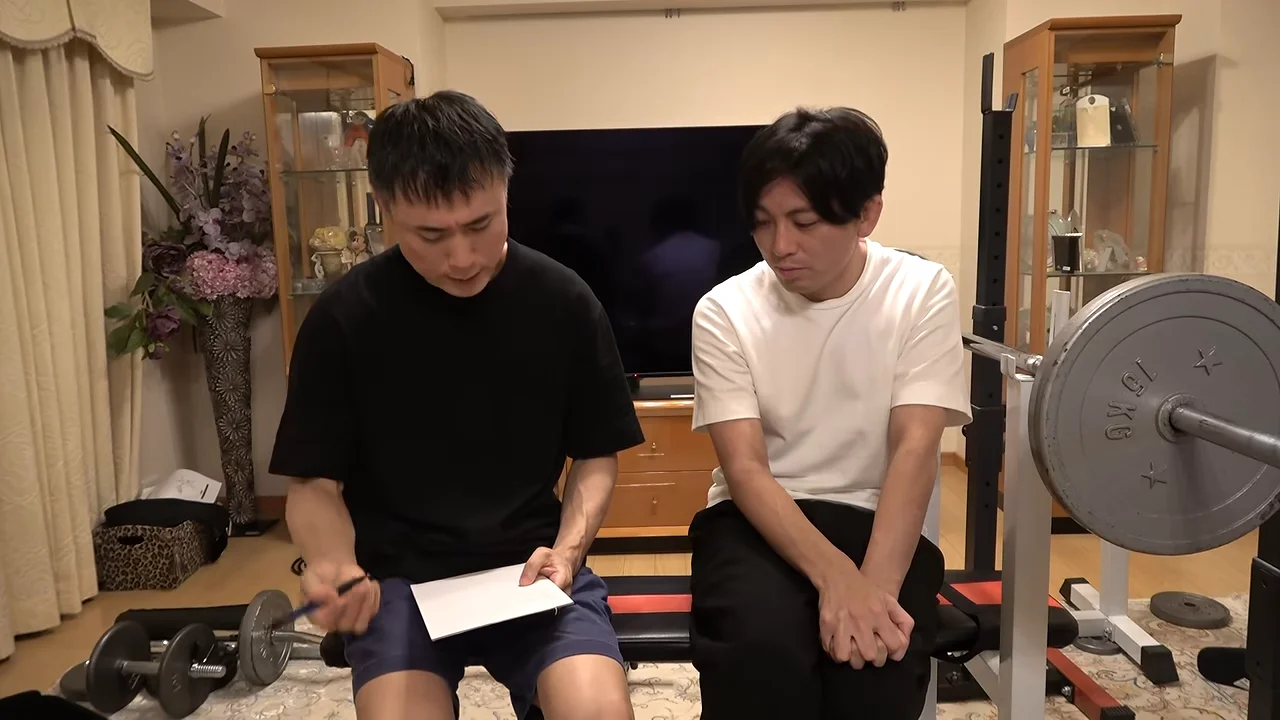 室内でトレーニング器具の前に座る2人の男性が、メモを見ながら話している。