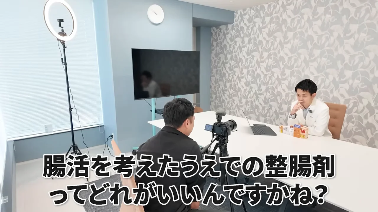 撮影カメラの前で白衣の男性が机に座り、画面下に大きな文字が表示されている。