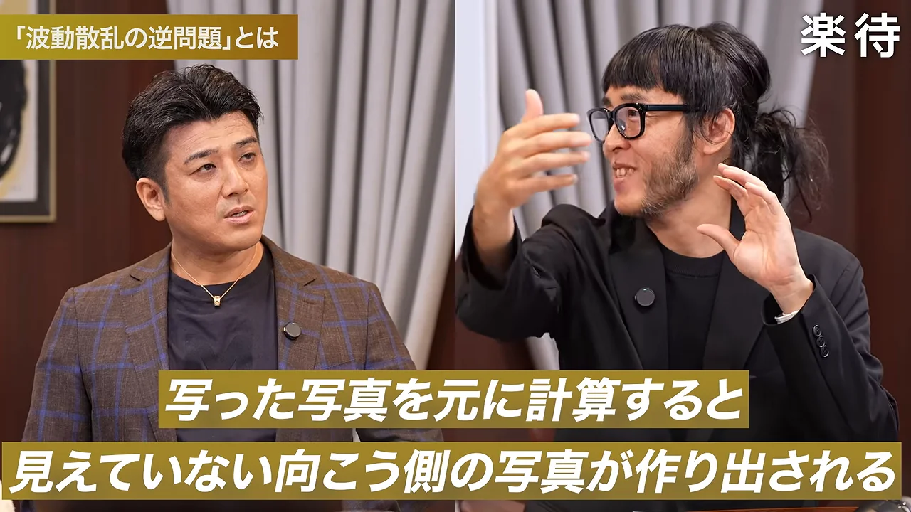 黒い服の男性が両手を動かして話し、隣の男性が見つめている。