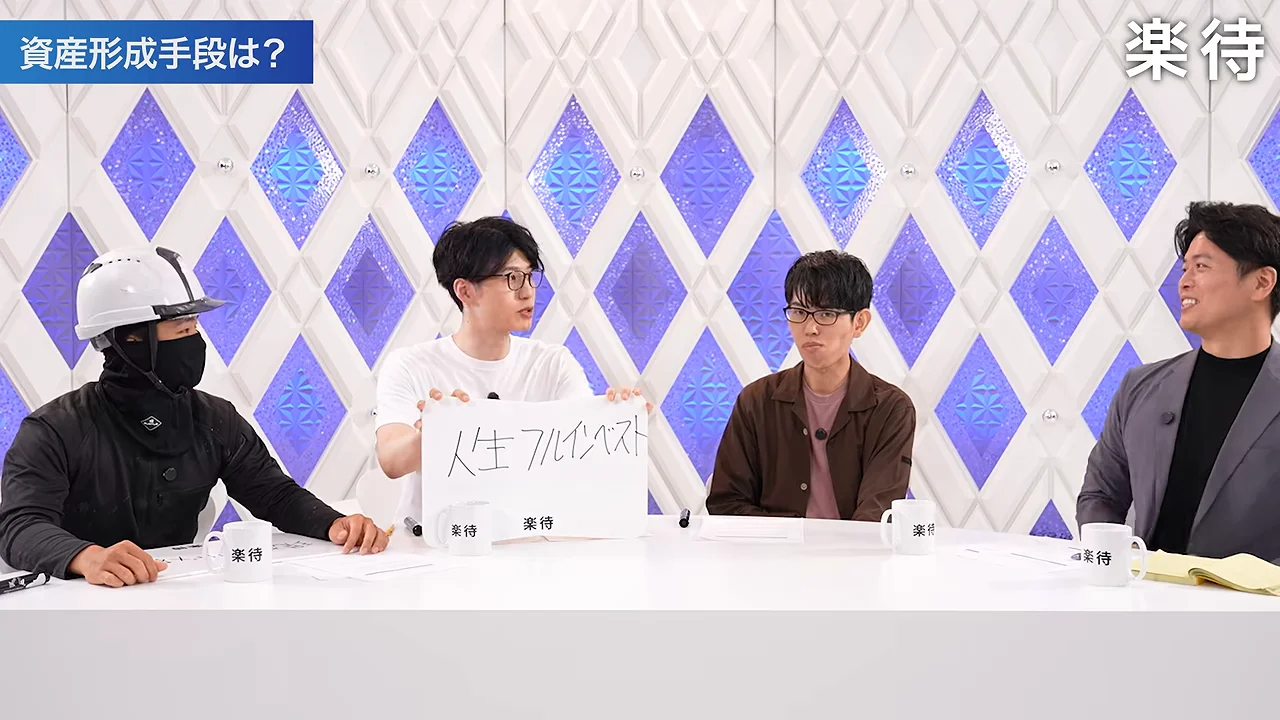 スタジオで4人の男性がテーブルを囲み、白いTシャツの男性がフリップを掲げています。