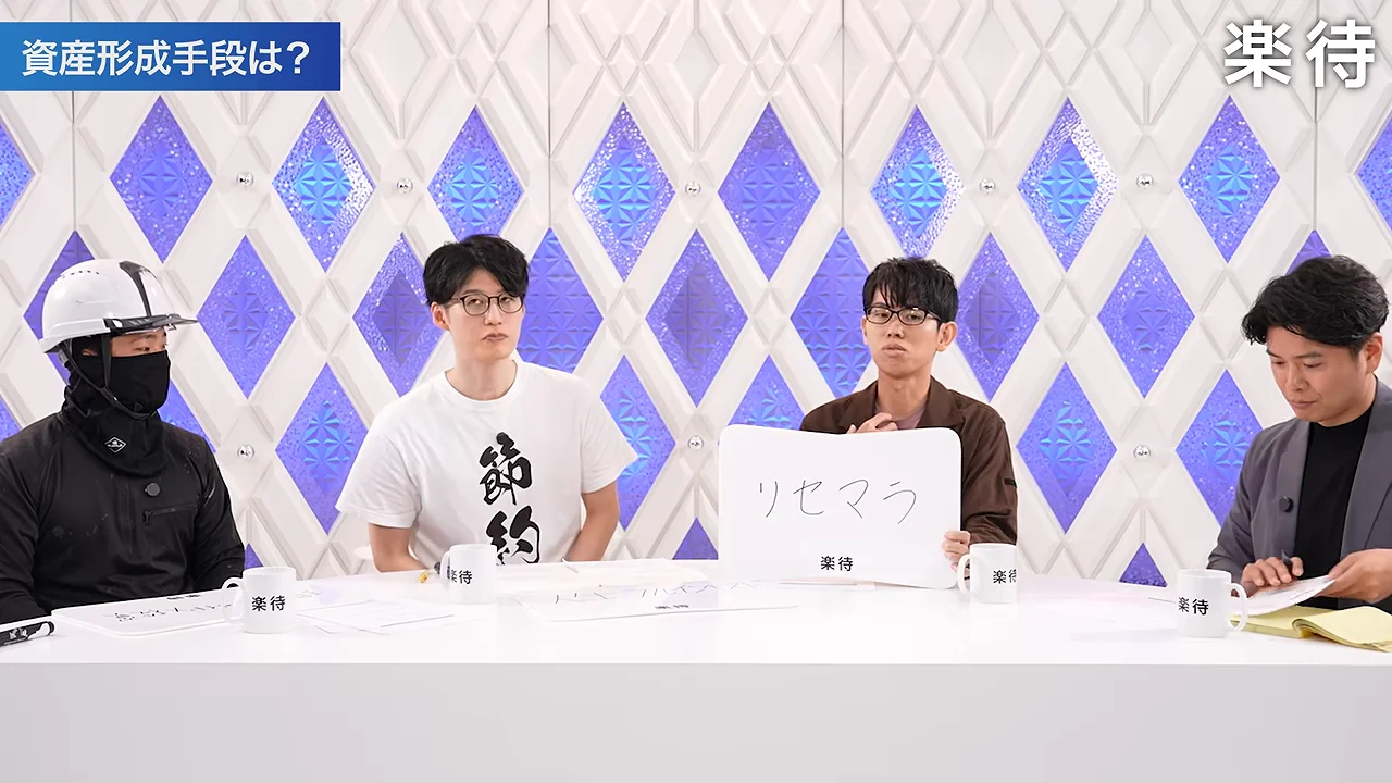 茶色のシャツを着た男性が「リセマラ」と書かれたフリップを掲げ、他の出演者がその様子を見つめています。