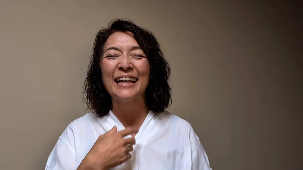 白い服を着た女性が胸元に手を添えて笑顔で話しています。