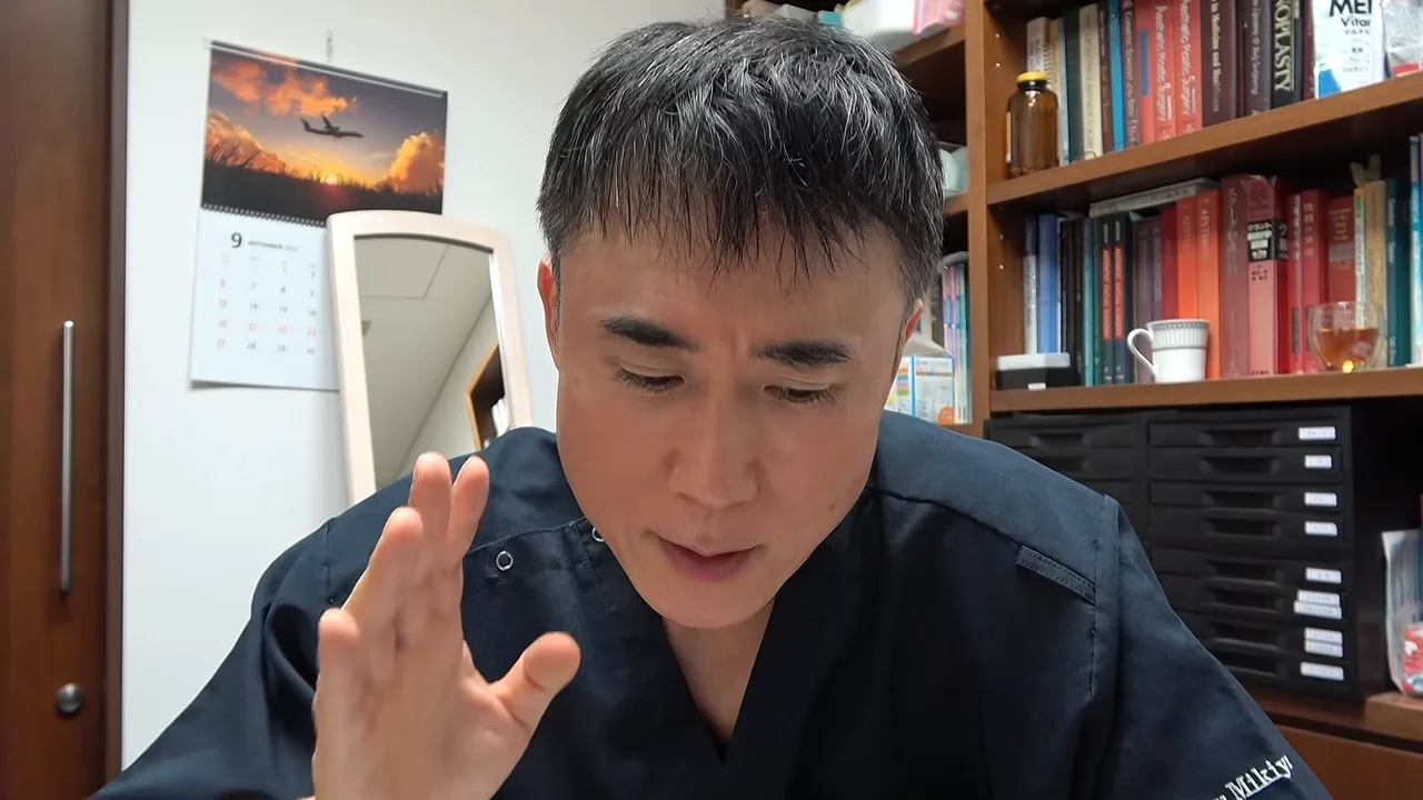 部屋の中で本棚を背にして右手でジェスチャーを交えながら話す男性