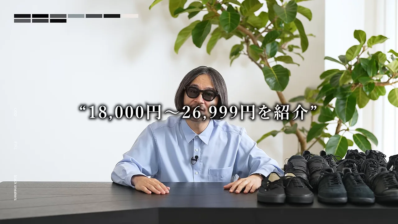 複数の黒いスニーカーが置かれたテーブルの前で、サングラスをかけた男性が座っています。
