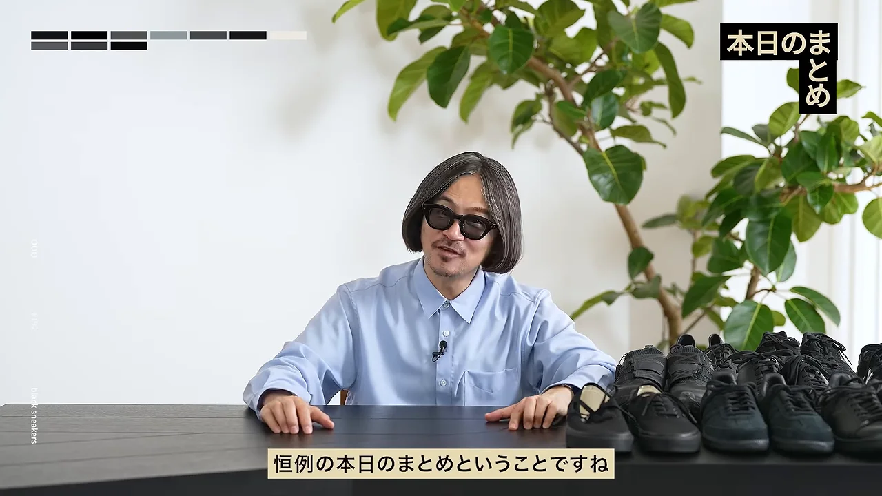 黒いスニーカーが並ぶテーブルの前に、サングラスをかけた男性が座っています。