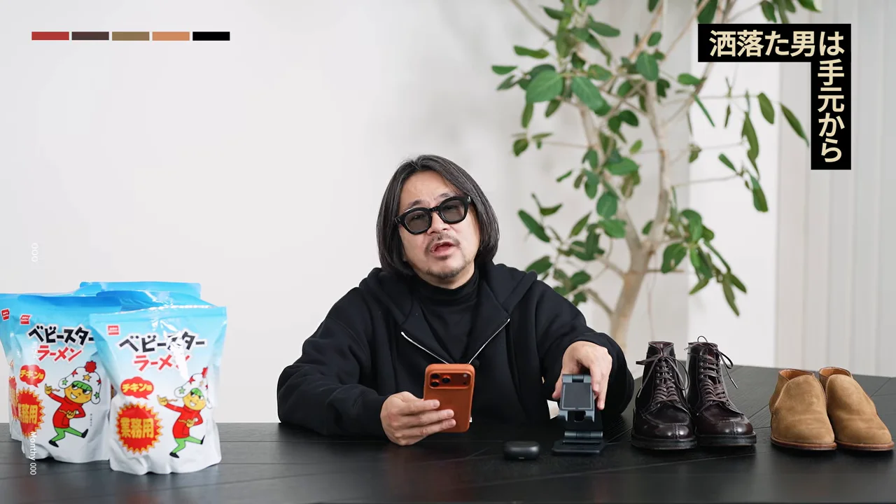 サングラスをかけた男性が、ベビースターラーメンや靴、スマートフォンが並ぶテーブルの前に座っている。