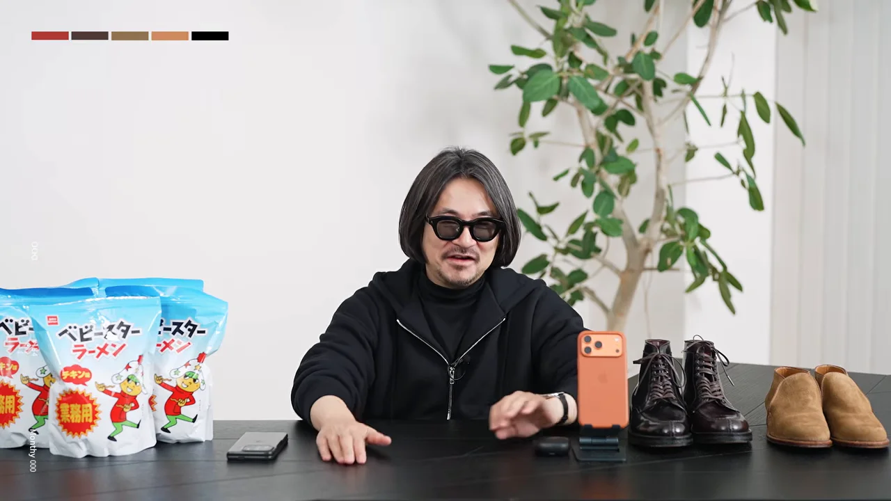 男性がテーブルの上に置かれた小物の前に両手を置き、前を向いて話している。