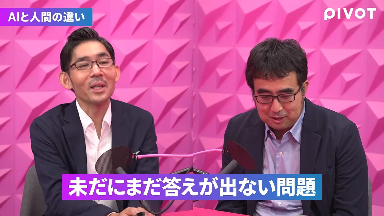 ピンクの幾何学模様の壁を背景に、スーツ姿の眼鏡をかけた2人の男性が座って話しています。