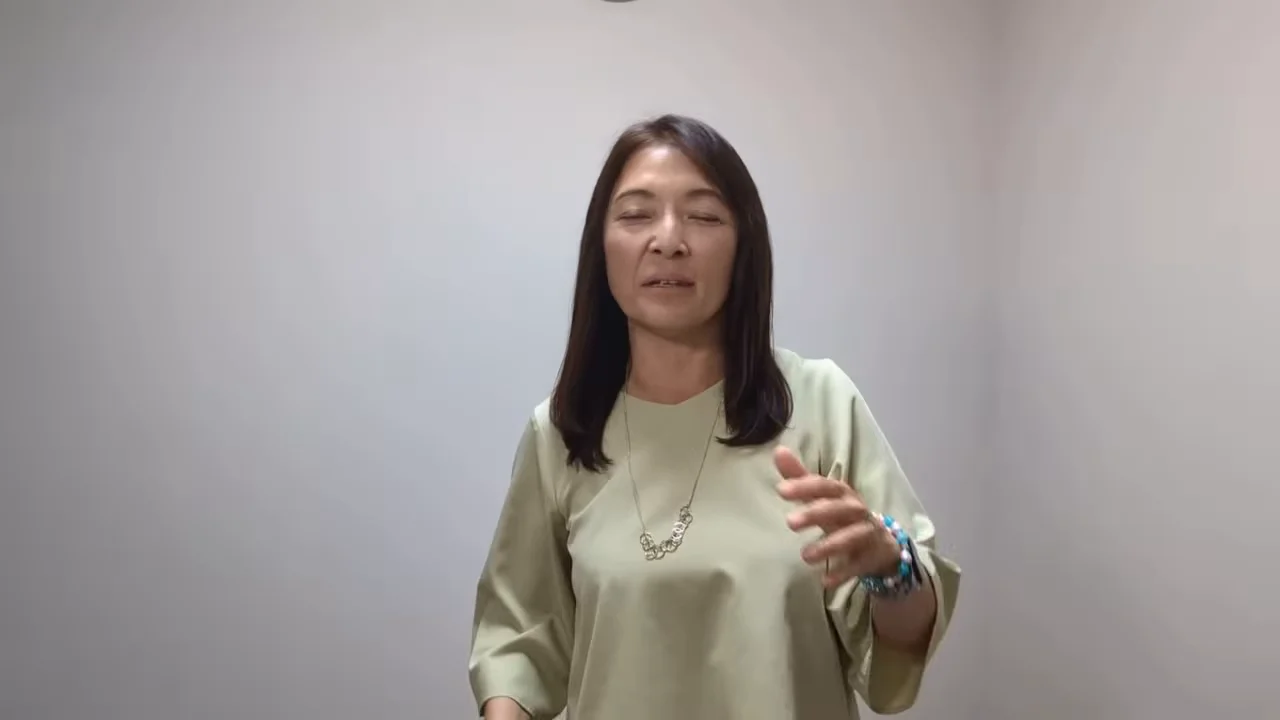 白い壁を背景に、左手を動かしながら話す女性