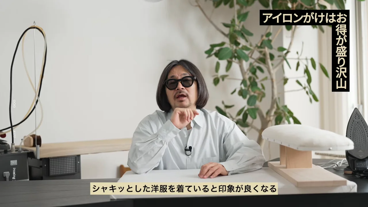 サングラスをかけた男性がアイロン台の置かれた机の前に座り、カメラに向かって話しています。