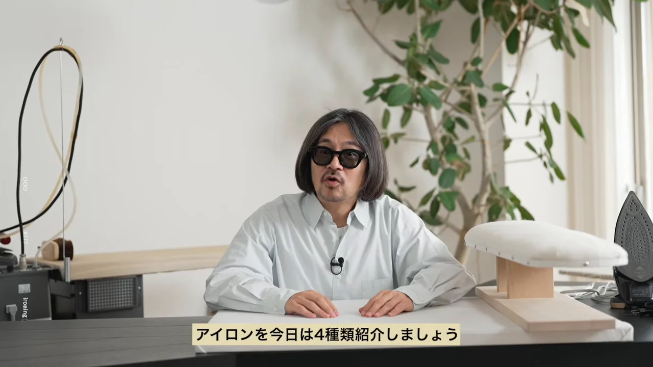 サングラスをかけた男性が机に両手を置いて座り、話しています。