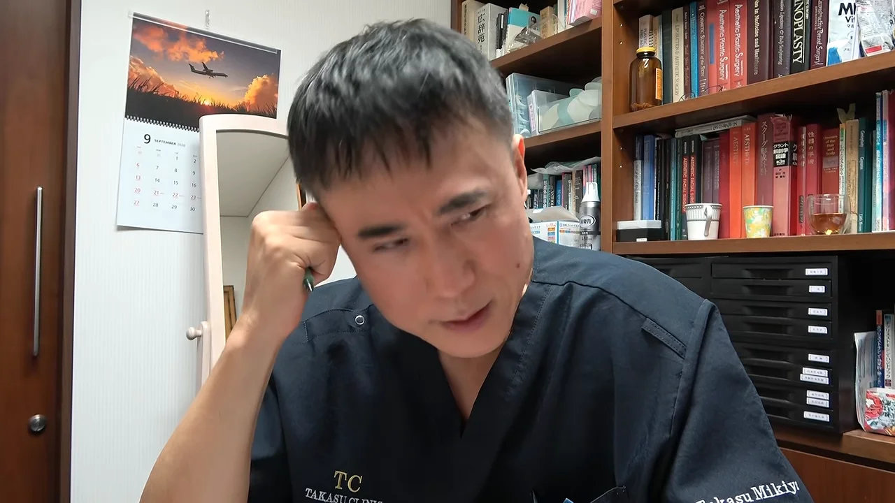 本棚を背にした男性が側頭部に手を当てて下を向いています。