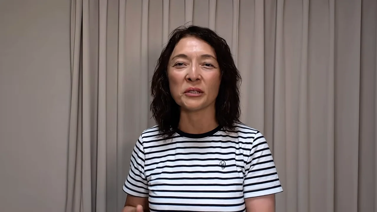 カーテンの前に立つボーダー柄のTシャツを着た女性がカメラに向かって話している様子。
