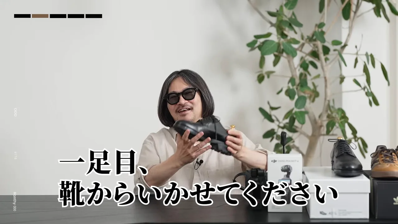 机の前に座った男性が、購入した黒い革靴を両手で持ち上げて紹介しています。
