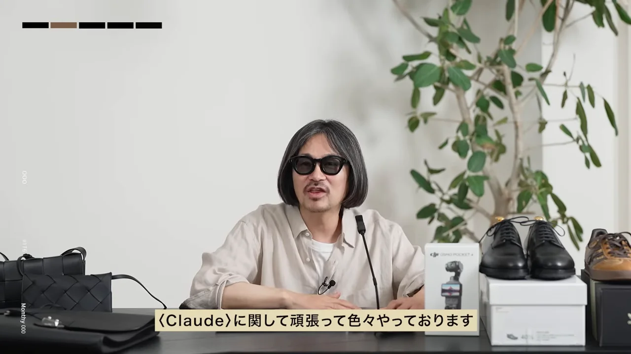 男性が机の上に靴やカバンなどの小物を並べ、正面を向いて話をしています。