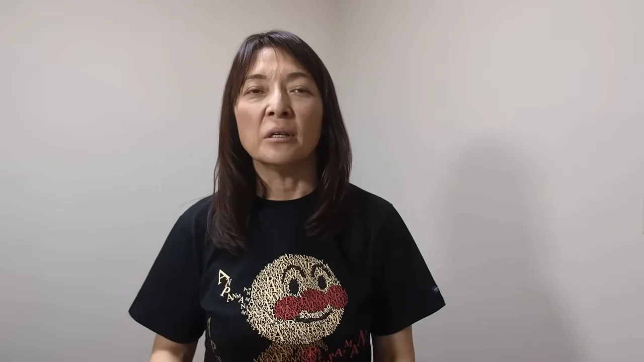 黒いTシャツを着た女性が白い壁の前に立ってカメラに向かって話している。