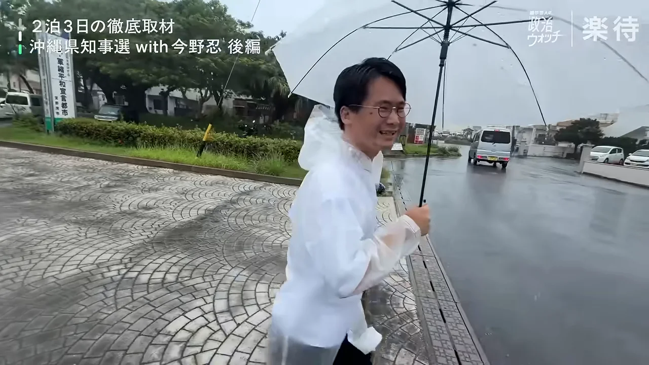 雨が降る屋外で、レインコートを着た眼鏡の男性が傘をさして歩いている様子。